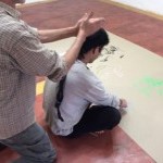 Image of a person sitting with legs crossed looking at a drawing placed on the floor with another person standing behind them.