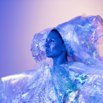 A performer in profile wears a voluminous, iridescent headpiece and blouse and is cast in a purple glow. 