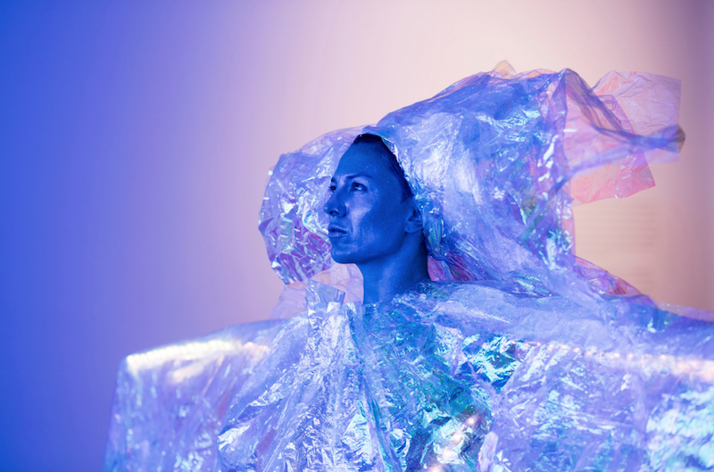 A performer in profile wears a voluminous, iridescent headpiece and blouse and is cast in a purple glow. 