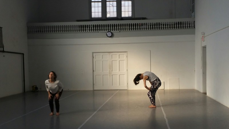 Two dancers in a sun-filled studio curve their backs. One is in side profile.