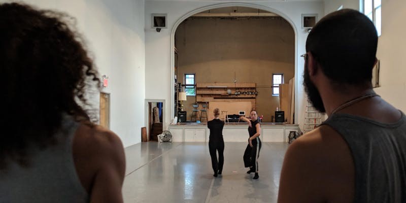 Performers are moving in front of an old gymnasium stage. The floor is grey and the walls are white.