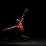 Male dancer in burgundy top and black tights sits in 4th position, back leg extended, facing stage left reaching in 4th.