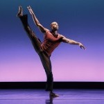 Male dancer in a side tilt, right leg high and arms extended in opposition, dressed in glittery burgundy top and black pants. 