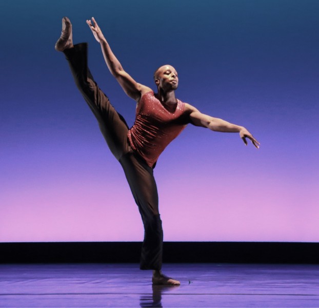 Male dancer in a side tilt, right leg high and arms extended in opposition, dressed in glittery burgundy top and black pants. 