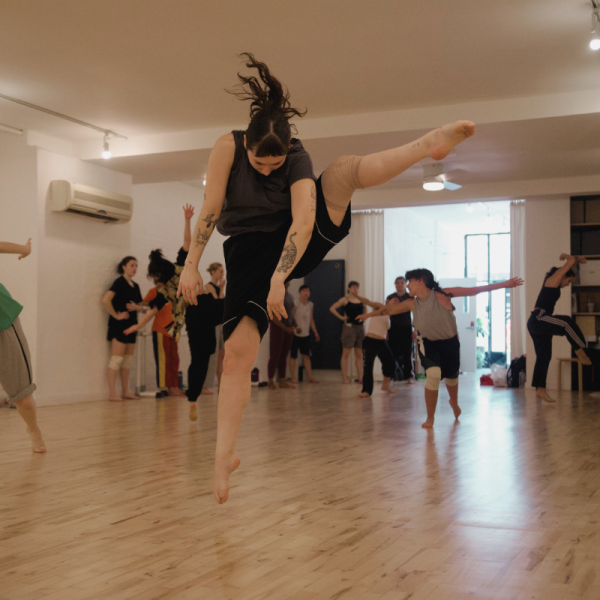 A dancer mid-air jumping in a dance studio with their left leg high and bent.