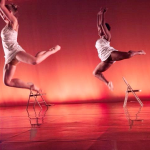 Two dancers leap through the air above chairs onstage during a contemporary dance performance