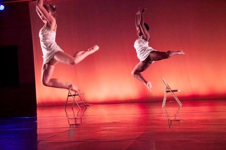 Two dancers leap above folding chairs on a stage lit with warm orange lighting