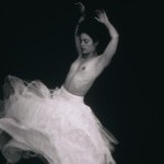 A black and white image of a woman swirling topless wearing a flowy white skirt