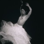 Black and white image of a woman swirling around topless in a flowy white skirt