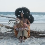 Two African American humans, one in a mask, another with big afro sits on a drift wood on a beach facing away from the water. 