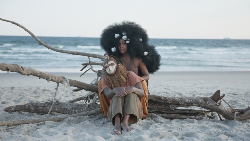 Two African American humans, one in a mask, another with big afro sits on a drift wood on a beach facing away from the water. 