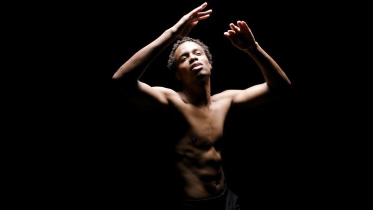 Photo of a man without his shirt on, standing in black abyss, lit from above