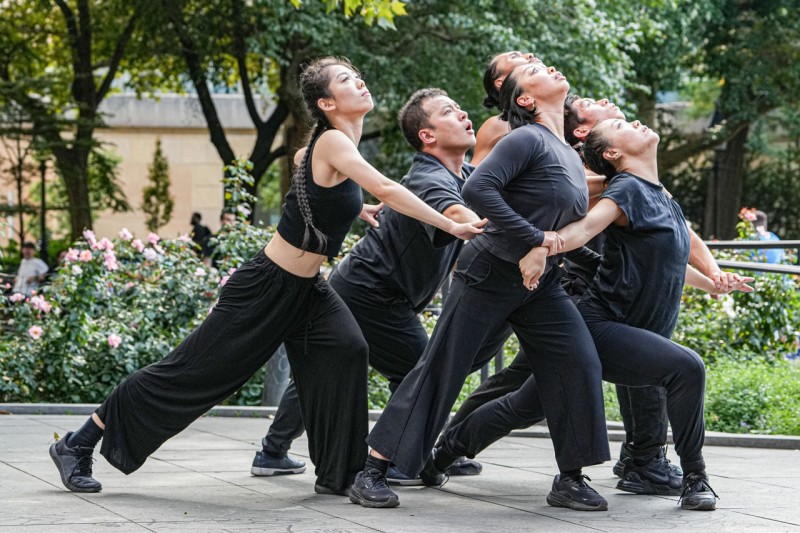 Dancers dressed in black, holding each other in a V formation, faces upward towards the sky