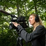 a photo of Founder Nel Shelby with video camera - she's standing in a wooded area and is wearing all black, with headphones.