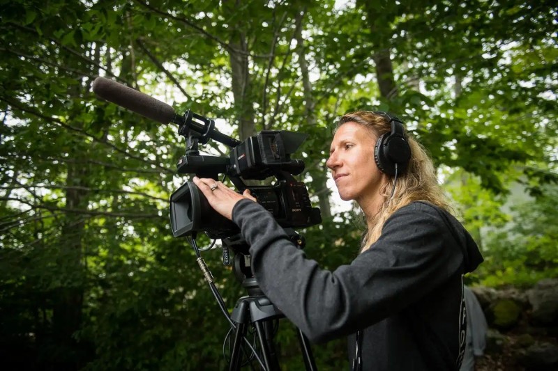 a photo of Founder Nel Shelby with video camera - she's standing in a wooded area and is wearing all black, with headphones.