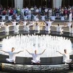 Many dancers, dressed completely in white, sit in a circle surrounding a fountain. 