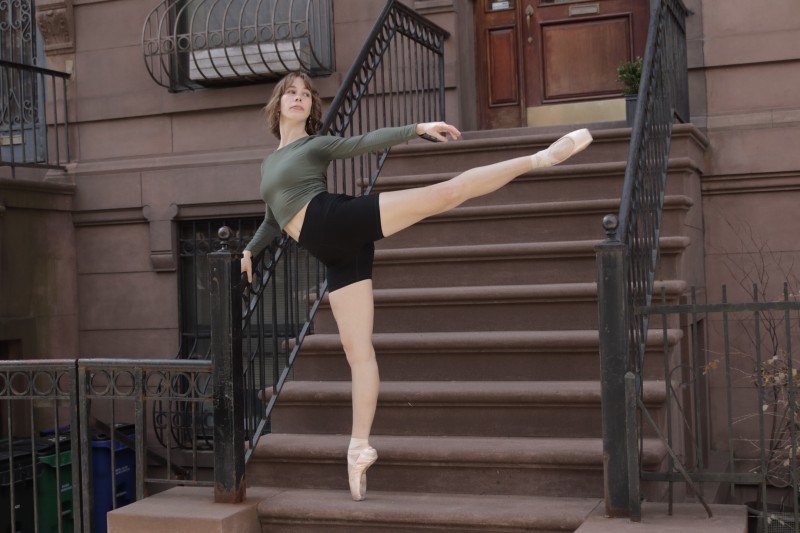 Full-body action shot of a female dancer in an arabesque in front of a NYC brownstone showcasing urban style