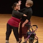 Vanessa (Night Sky) Cruz and Bailey Anderson stand on hardwood flooring, Bailey's rollator behind, with geometric arms