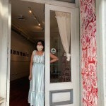 A woman in a long blue sundress stands in an open doorway, welcoming guests to the gallery.