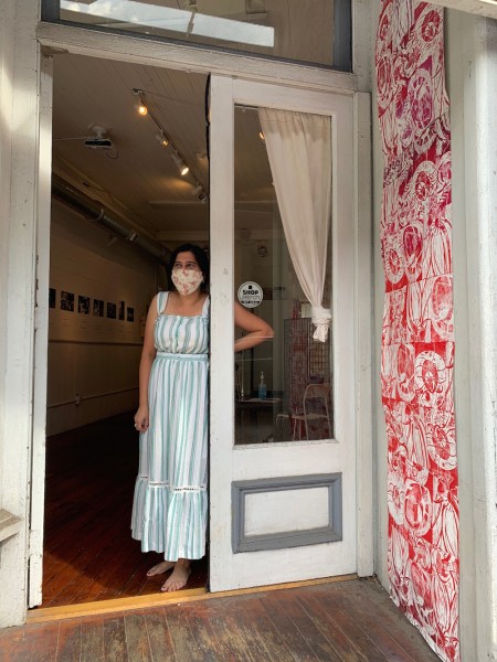 A woman in a long blue sundress stands in an open doorway, welcoming guests to the gallery.