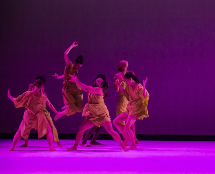 Seven dancers on stage dancing in green costumes under purple lighting