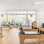 a white studio with wooden floors and with various pilates and gyrotonic equipment. windows in the back 