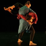 Two dancers perform a lift on stage. One is draped across the other's stomach with legs extended and face leaning downwards.