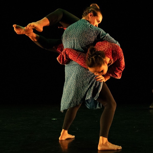 Two dancers perform a lift on stage. One is draped across the other's stomach with legs extended and face leaning downwards.