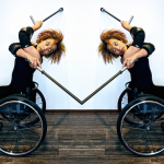 Alice Sheppard seated in her wheelchair with two crutches, crosses her body and arches her spine to look behind.