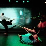 Two dancers in garish costumes pull a rope, suspending a third dancer in an off-balance attitude derriere