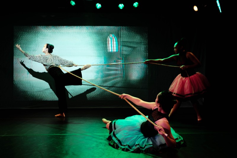 Two dancers in garish costumes pull a rope, suspending a third dancer in an off-balance attitude derriere