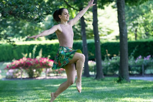 A dancer, dressed in a brown leotard and green skirt, skips through a grassy field.