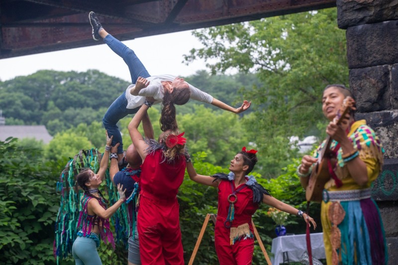 4 Dancers lifting one dancer into the air as a musician plays and sings in a colorful outdoor performance