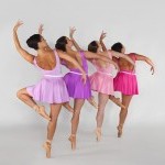 Four dancers in various shades of pink in pointe shoes on a diagonal in a retire position facing away from the camera.