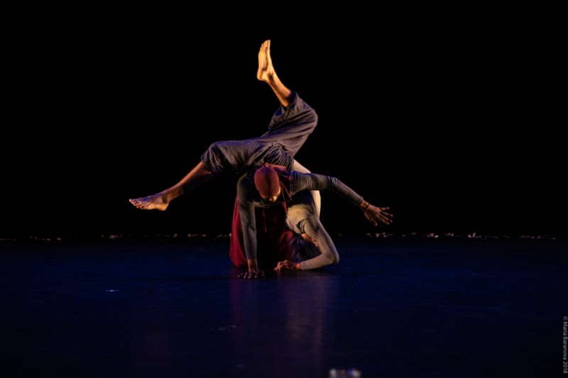 Two performers in a black box, one on knees and legs and the other dancer rolls on their back with legs in the air.