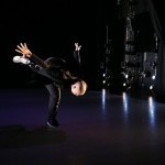 A dancer wearing all black in a black box theater falling towards the ground with one leg on the floor with the other in the air