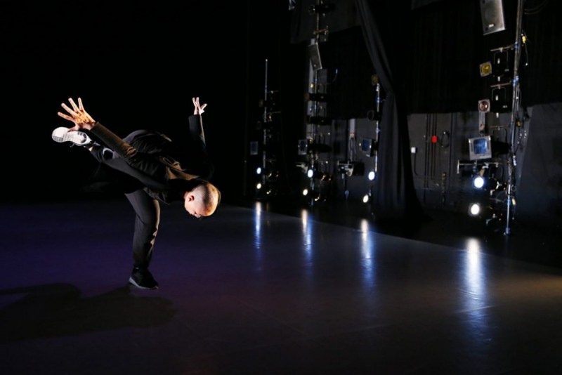 A dancer wearing all black in a black box theater falling towards the ground with one leg on the floor with the other in the air