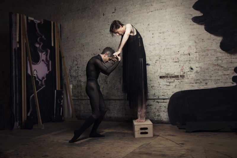 Two people both in black. One person on a cinder block reaching out their arms to the person below standing on the floor.