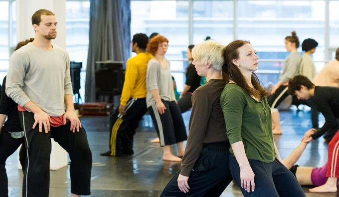 Dancers in a studio in pairs leaning against each others backs doing partnering work.