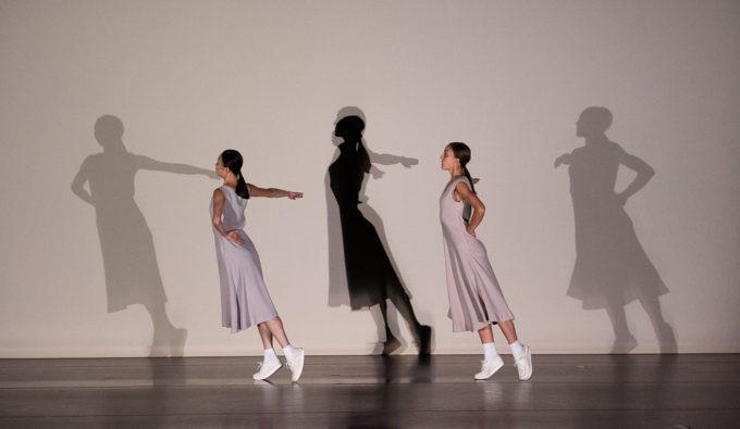 Two women in basic light grey dresses in their dance positions with the shadows projected behind them.