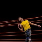 A woman gracefully bending her back in front of a black backgroup with multiple orange lines.