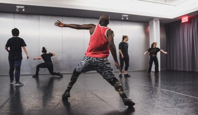 Dancers in a studio, with the focus on a person facing the wall in a lung with his arm at to the left in motion.