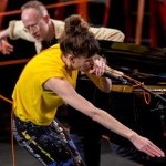 Joanna Kotze in a bright yellow tank top and multi-colored legginging with an arched back, extended arm and microphone in hand.