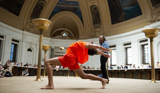 A person in a red/orange outfit lunging with the back bent while another person stands behind them in a pantheon