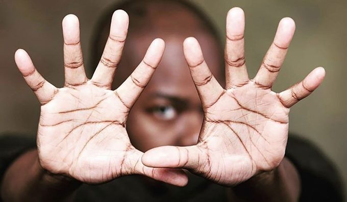 Jaamil Olawale Kosoko reaching the palms of his hands out to the point where they are almost covering his face.