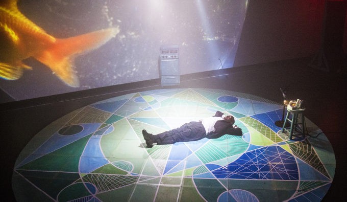 A person of color in a suit lying on the ground of a stage. On the black stage is a circle with earth tones in patterns.