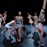 Eight dances moving in and out of motion in a black box wearing neutral colors like grays and whites.