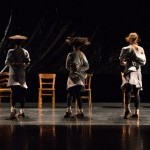 Three dancers is muted colors like a soft grey and black, turning in motion with wooden chairs behind them.
