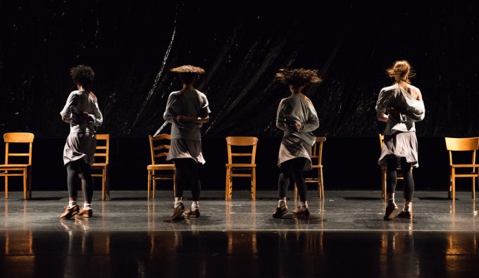 Three dancers is muted colors like a soft grey and black, turning in motion with wooden chairs behind them.