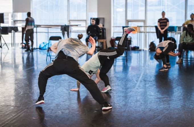 Two dancers with their backs to the views dance in the studio, one with the hands and leg on the ground with another leg up.